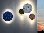 Four Clérachic round wall lights in blue, black, white and pink terrazzo glow on a ribbed metal wall at dusk.