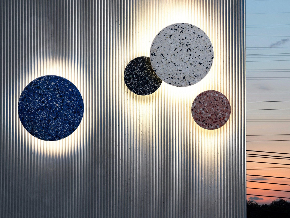 Four Clérachic round wall lights in blue, black, white and pink terrazzo glow on a ribbed metal wall at dusk.