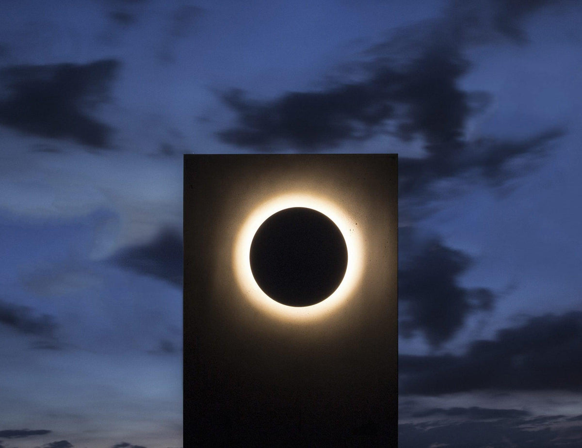 A plain black Clérachic disc wall light on a black panel against a dusky blue sky, ringed with warm light.