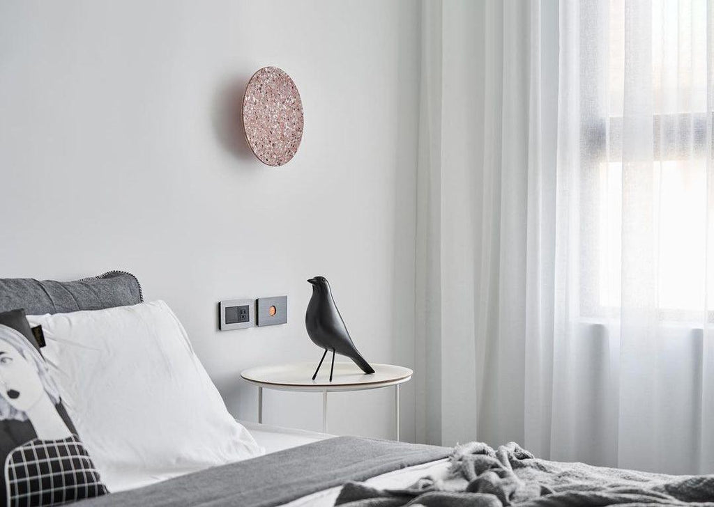 A pink terrazzo Clérachic round wall light hangs above a bed with white linen, beside sheer curtains and a side table.