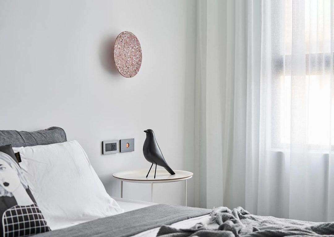 A pink terrazzo Clérachic round wall light hangs above a bed with white linen, beside sheer curtains and a side table.