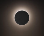 A plain black Clérachic round wall light, its edge ringed with a soft white halo on a dark wall.
