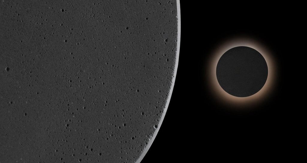 Close view of the pitted dark cement surface of this Clérachic round wall light, with a second one glowing beyond it.