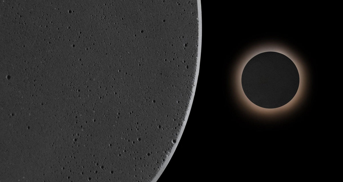 Close view of the pitted dark cement surface of this Clérachic round wall light, with a second one glowing beyond it.
