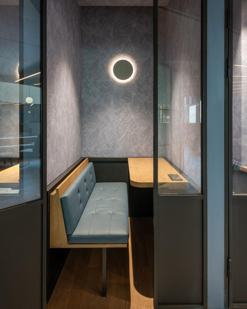 A Clérachic round wall light glows above a small meeting booth with a blue-grey bench seat, behind a glass partition.