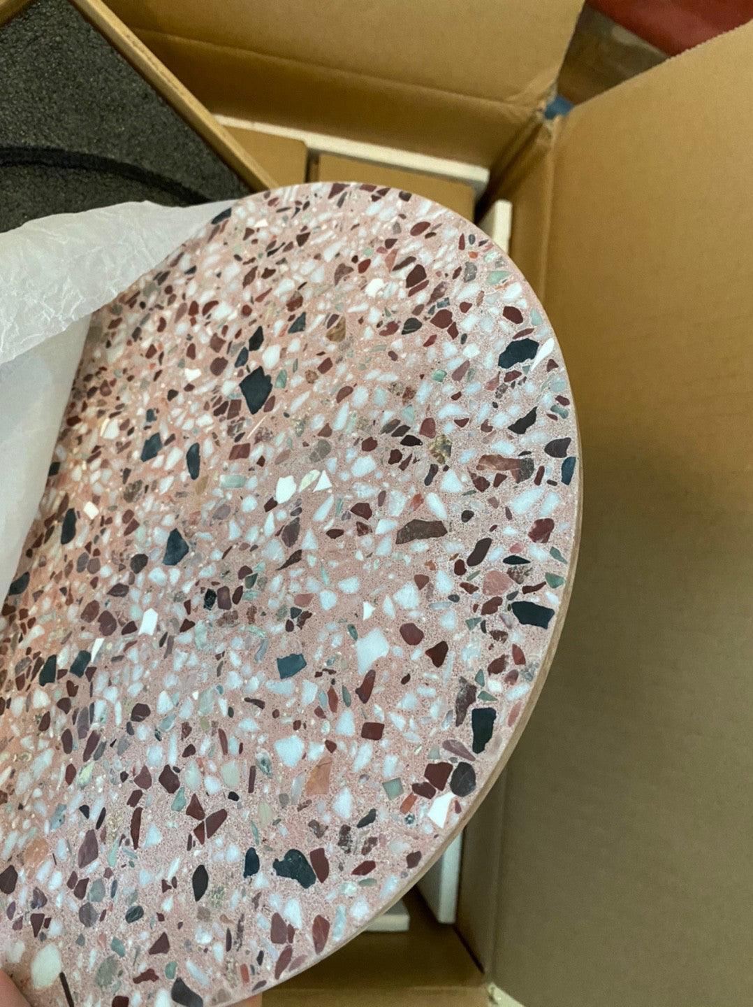 A pink terrazzo Clérachic round wall light, with a pale lining and dark chips, lying in an open cardboard box.