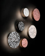 A cluster of white, black and pink terrazzo Clérachic round wall lights, lit warmly on a very dark wall.