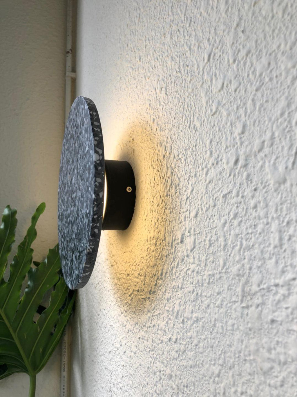 Side view of a terrazzo Clérachic round wall light standing off a textured white wall on its black mount, glowing warm.