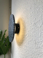 Side view of a terrazzo Clérachic round wall light standing off a textured white wall on its black mount, glowing warm.