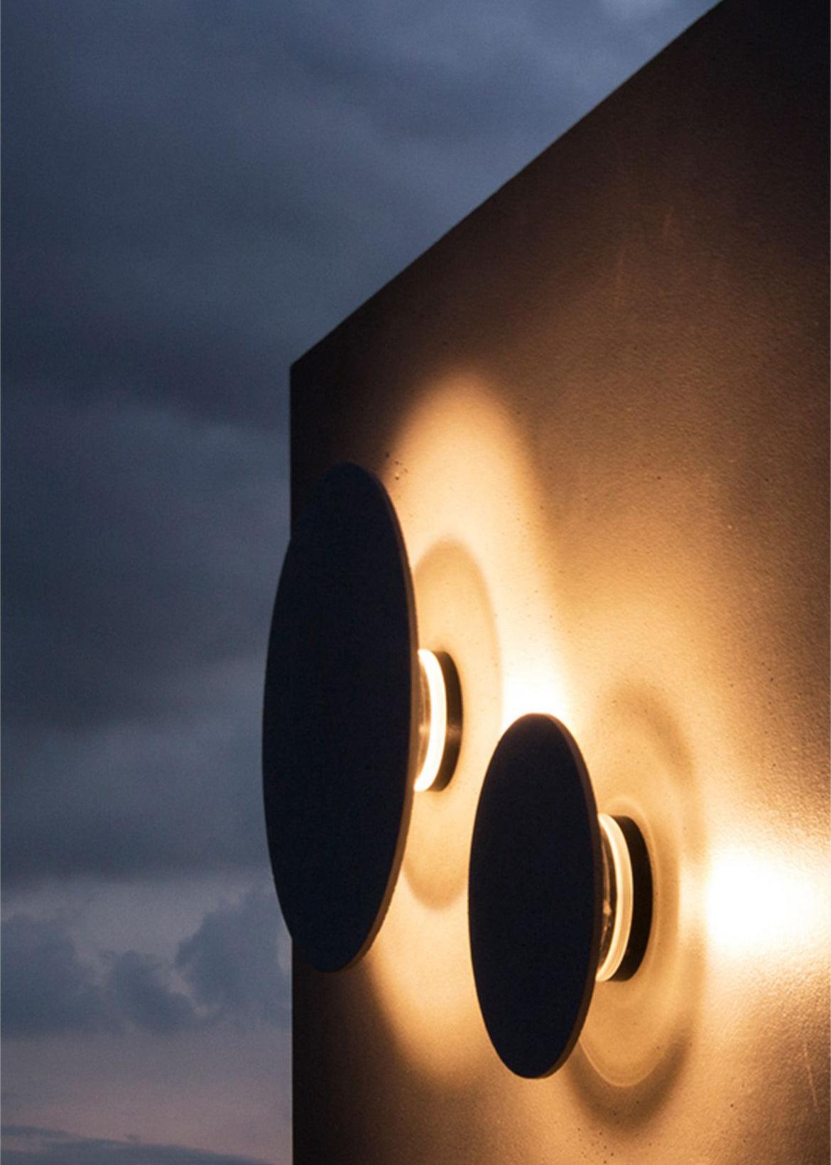 Two plain black Clérachic round wall lights with brass rings glow warmly against a dark panel and a dusky sky.