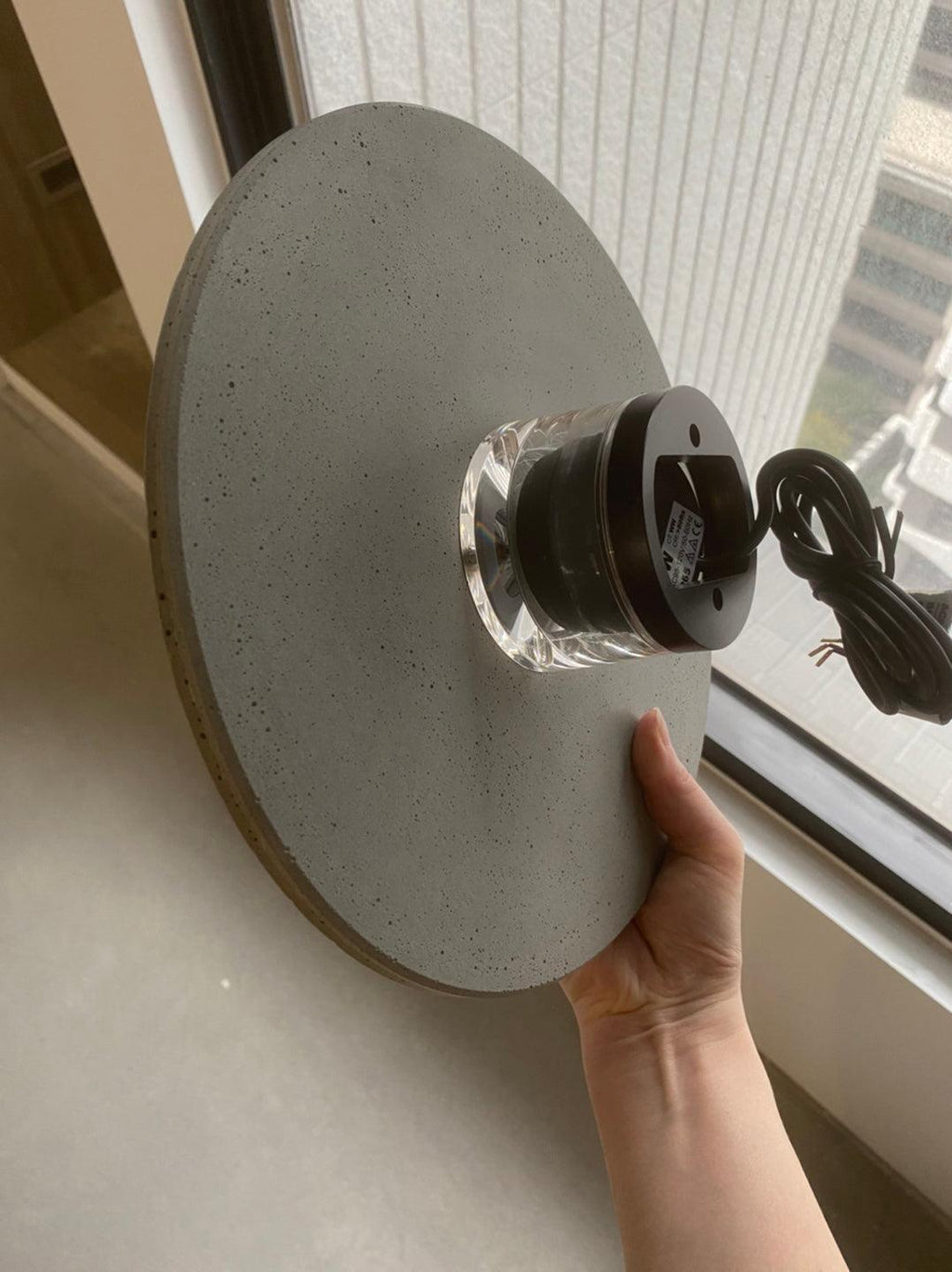 A hand holds the back of a cement-grey Clérachic round wall light, showing its clear mounting block and cable.