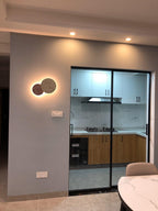 Two small Clérachic round wall lights glow beside a sliding glass kitchen door in a grey-walled room.
