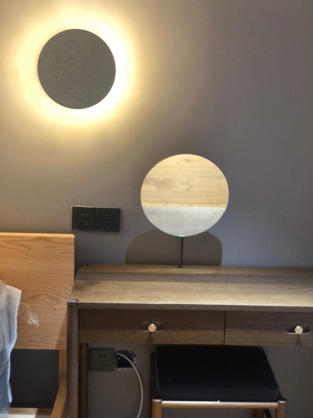 A grey Clérachic round wall light glows above a wooden desk with a round mirror, on a grey wall.