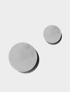 Two cement-grey Clérachic round wall lights in different sizes, with soft shadows, on a pale grey backdrop.