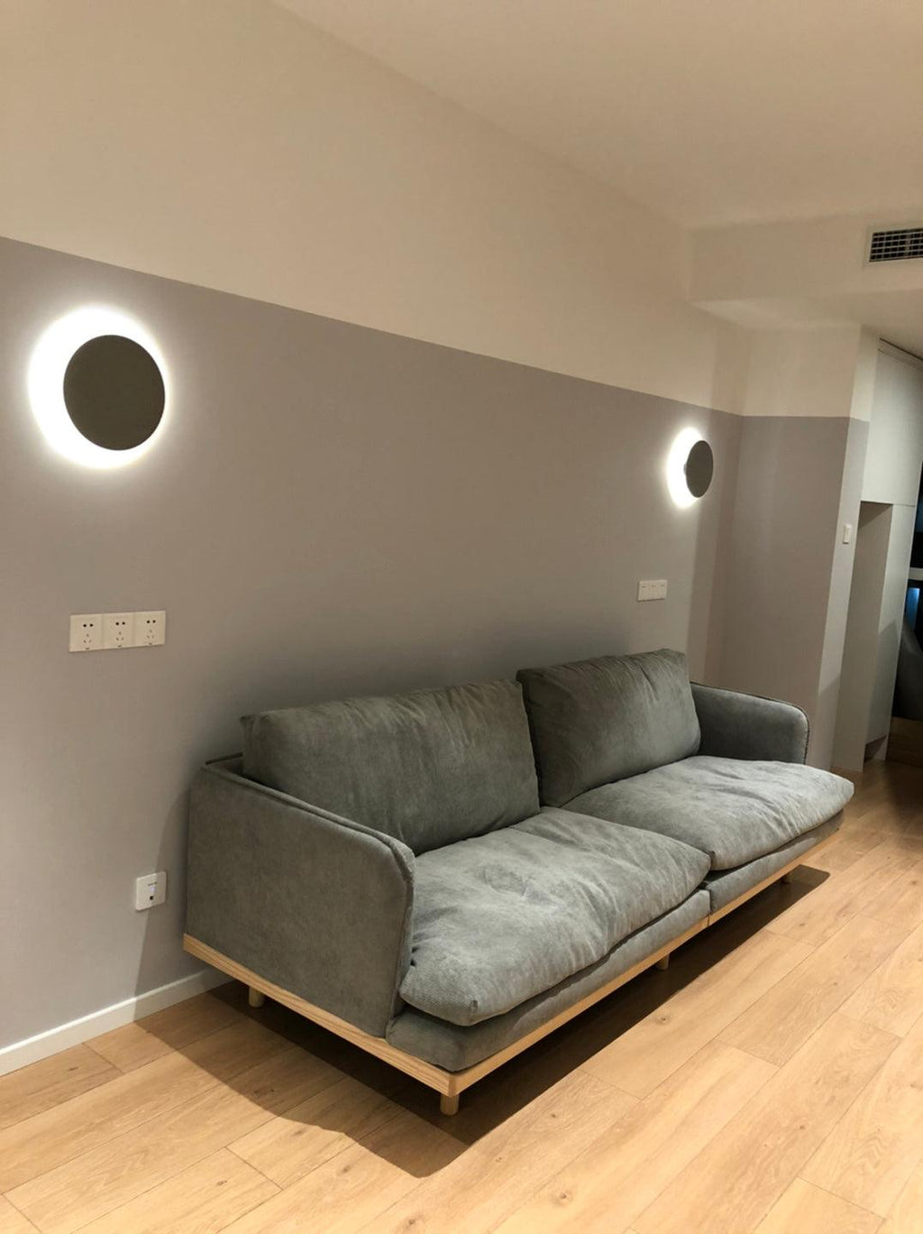 A Clérachic round wall light glows beside a second one in a lounge with a grey sofa and pale wood floor.