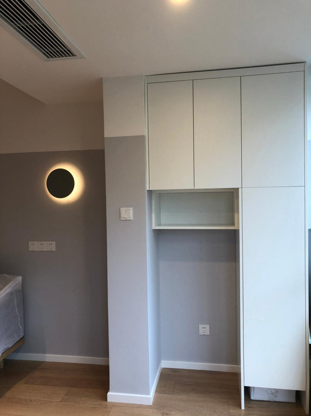 A plain black Clérachic round wall light glows on a grey wall beside tall white cupboards.