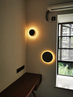 Two plain black Clérachic round wall lights glow warmly in a corner by a black-framed window, above a wooden desk.
