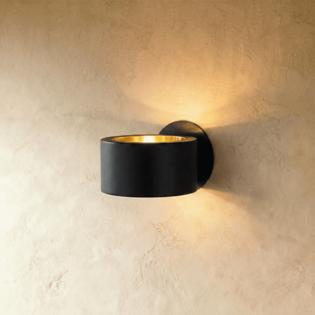 Clérachic round black metal wall lamp with a gold-lined top opening, casting warm light up the limewashed wall behind it.