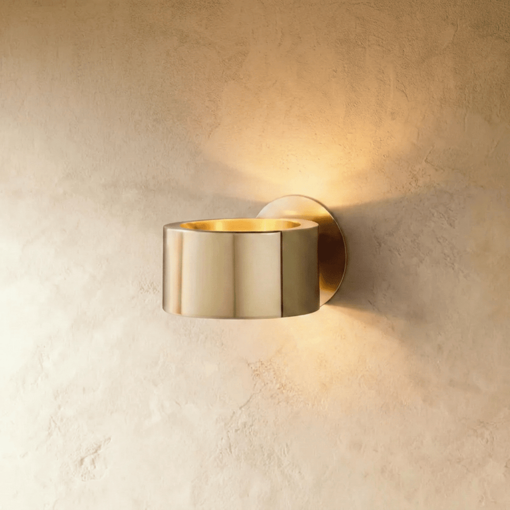 Clérachic round gold metal wall lamp with a polished drum body and glowing gold-lined top, on a soft limewashed wall.