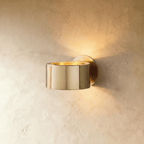 Clérachic round gold metal wall lamp with a polished drum body and glowing gold-lined top, on a soft limewashed wall.