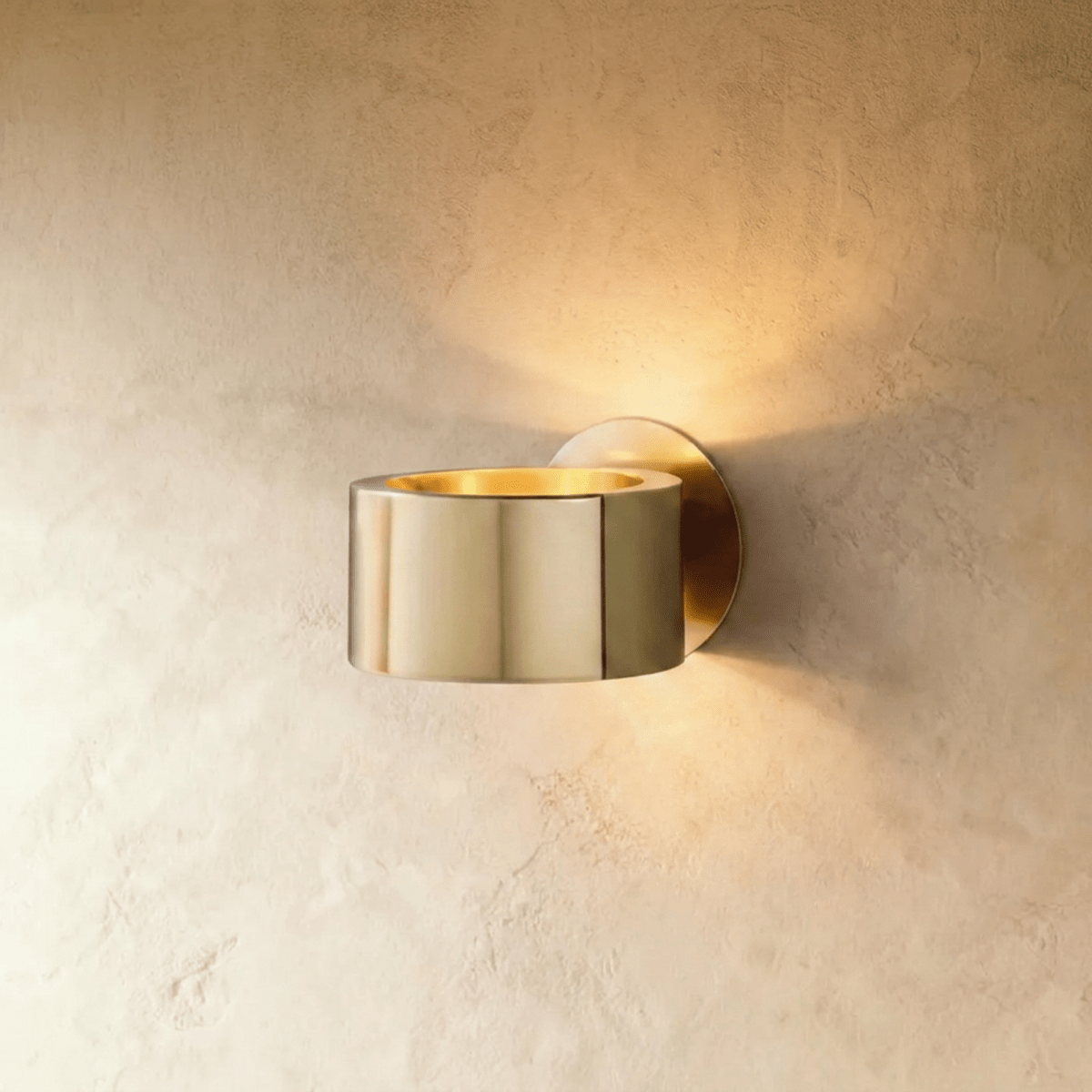 Clérachic round gold metal wall lamp with a polished drum body and glowing gold-lined top, on a soft limewashed wall.