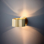Clérachic polished gold drum wall lamp throwing warm light up and down a slate grey wall in a fan-shaped glow.