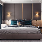 Clérachic pair of gold drum wall lamps glowing either side of a headboard on grey panelled bedroom wall above teal cushions.