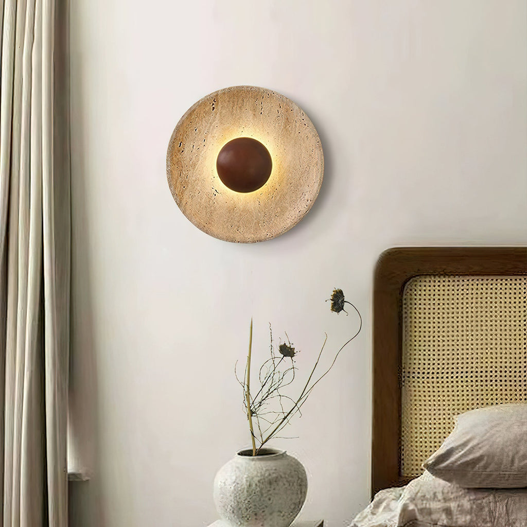 A lit Clérachic travertine wall light with a walnut dome, on a white wall beside a curtain, a rattan bedhead and a vase of dried stems.