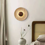 A lit Clérachic travertine wall light with a walnut dome, on a white wall beside a curtain, a rattan bedhead and a vase of dried stems.