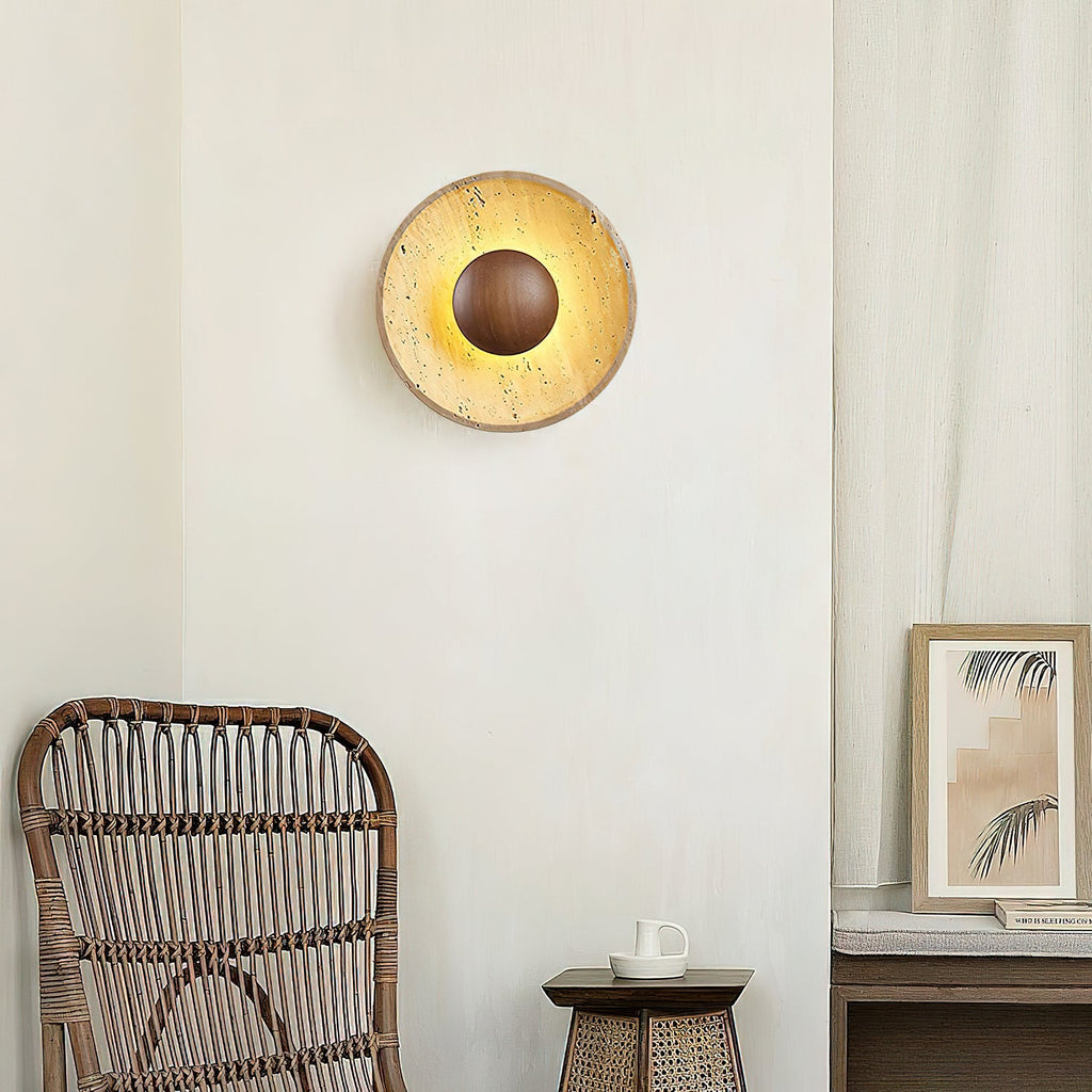 Clérachic round wall light in yellow travertine with a walnut centre dome, glowing above a rattan chair and a small framed print.