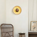 Clérachic round wall light in yellow travertine with a walnut centre dome, glowing above a rattan chair and a small framed print.