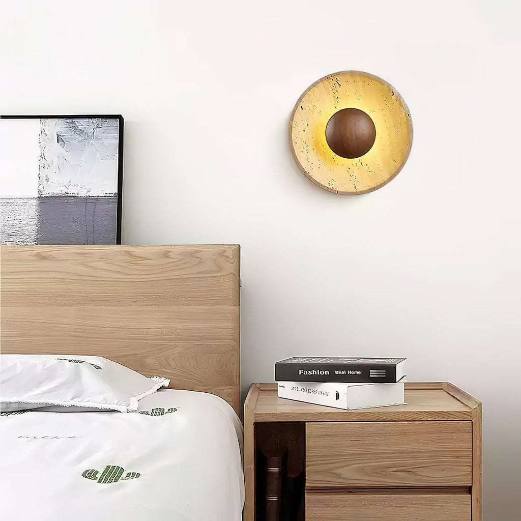 This Clérachic round travertine wall light glows above a pale wood bed and a bedside table holding two stacked books.