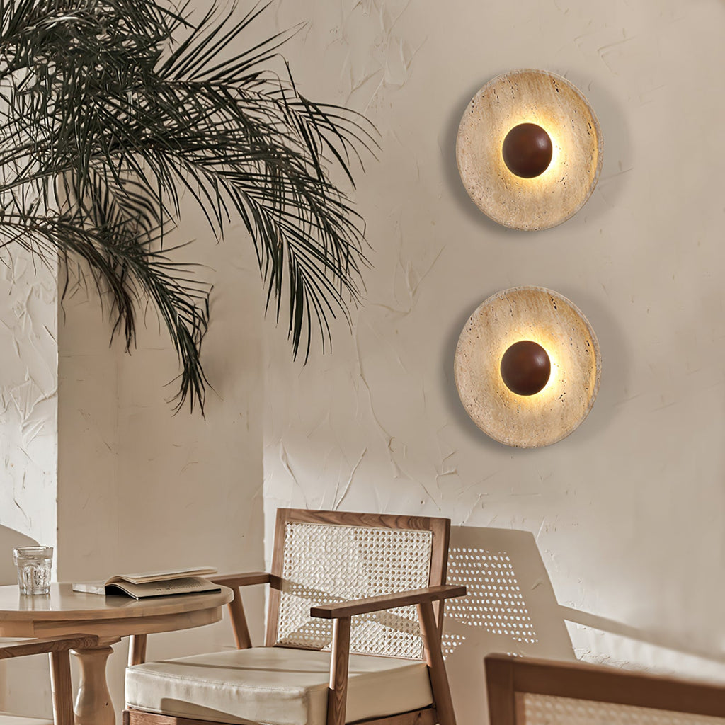 Two Clérachic travertine wall lights with walnut domes on a warm plaster wall, beside a palm and a rattan-backed dining chair.