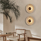 Two Clérachic travertine wall lights with walnut domes on a warm plaster wall, beside a palm and a rattan-backed dining chair.