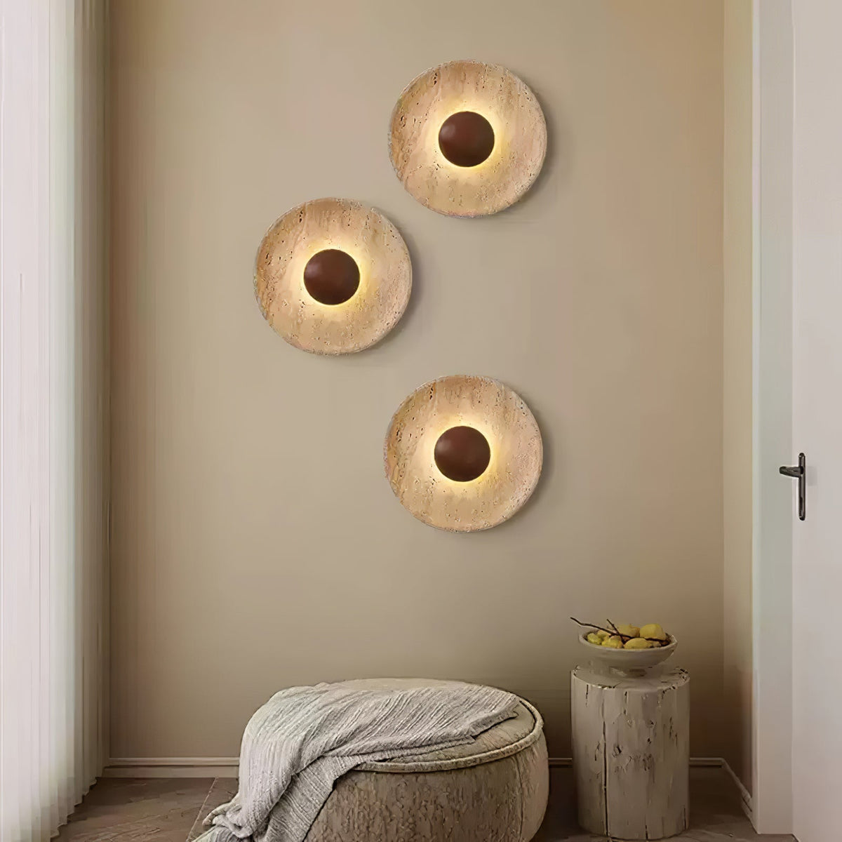 Three lit Clérachic round wall lights in travertine with walnut domes, set in a staggered group above a woven pouffe in a beige room.