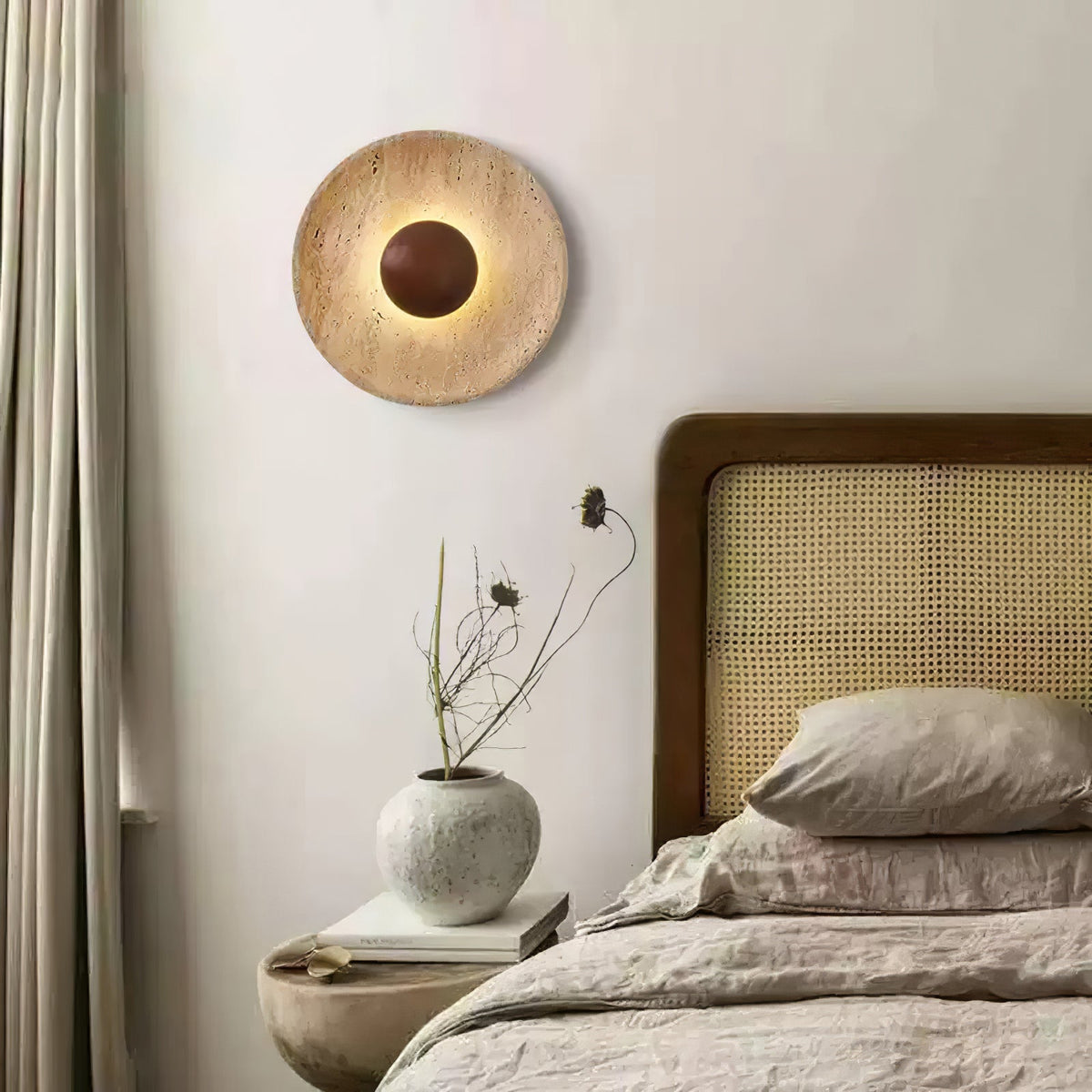 Clérachic travertine wall light with a walnut dome, lit, on a pale wall beside a curtain, above a linen bed and a round stone bedside table.