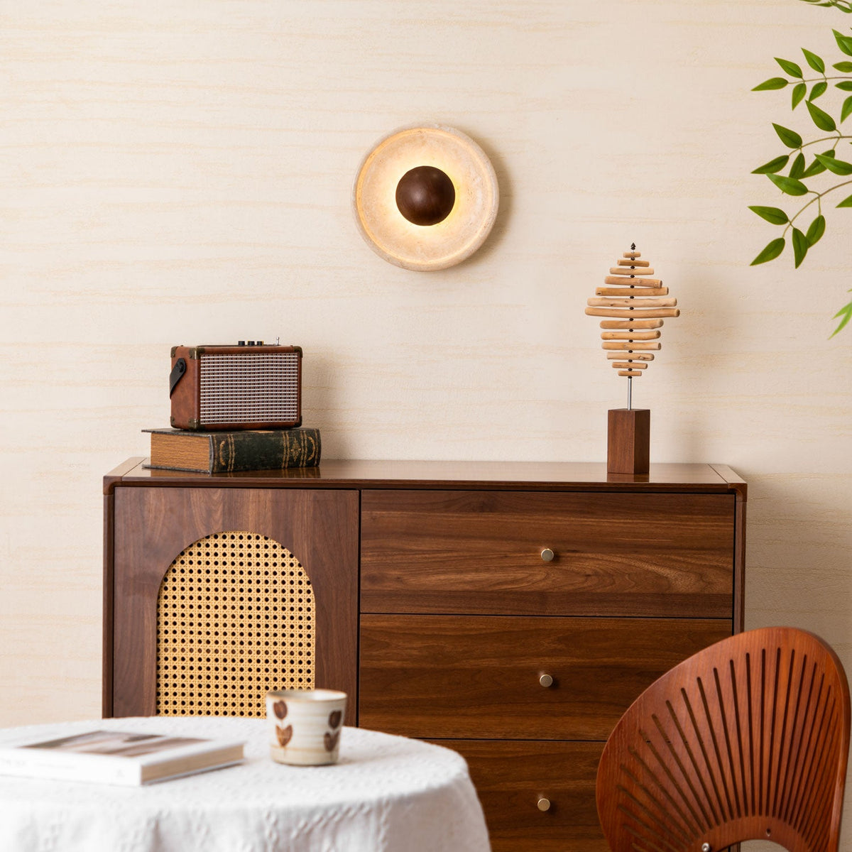 Lit Clérachic travertine wall light with a walnut dome on a cream wall, above a walnut cabinet with a rattan door and a retro radio.
