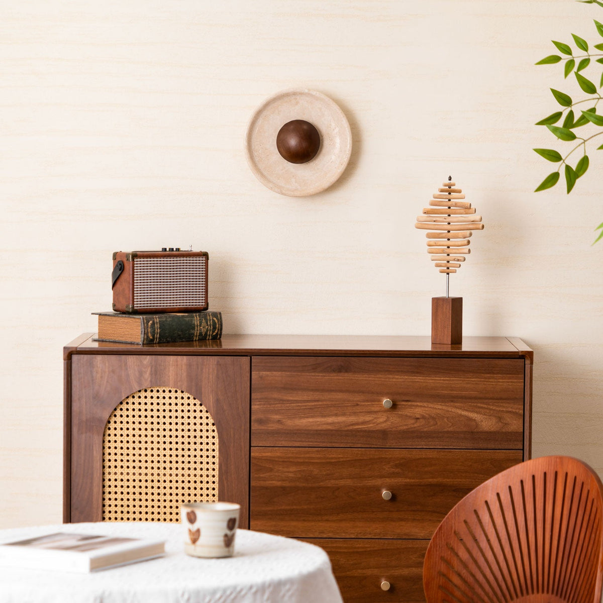 Unlit, a paler travertine version of this Clérachic wall light sits above a walnut cabinet with a rattan panel, a radio and a wooden sculpture.