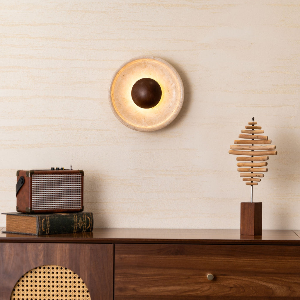 Lit Clérachic travertine wall light with a walnut dome glowing warm on a cream wall above a walnut cabinet, radio and wooden sculpture.