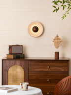 This Clérachic wall light lit warm on a cream wall, above a walnut cabinet with a radio, a stepped wood sculpture and a cup.