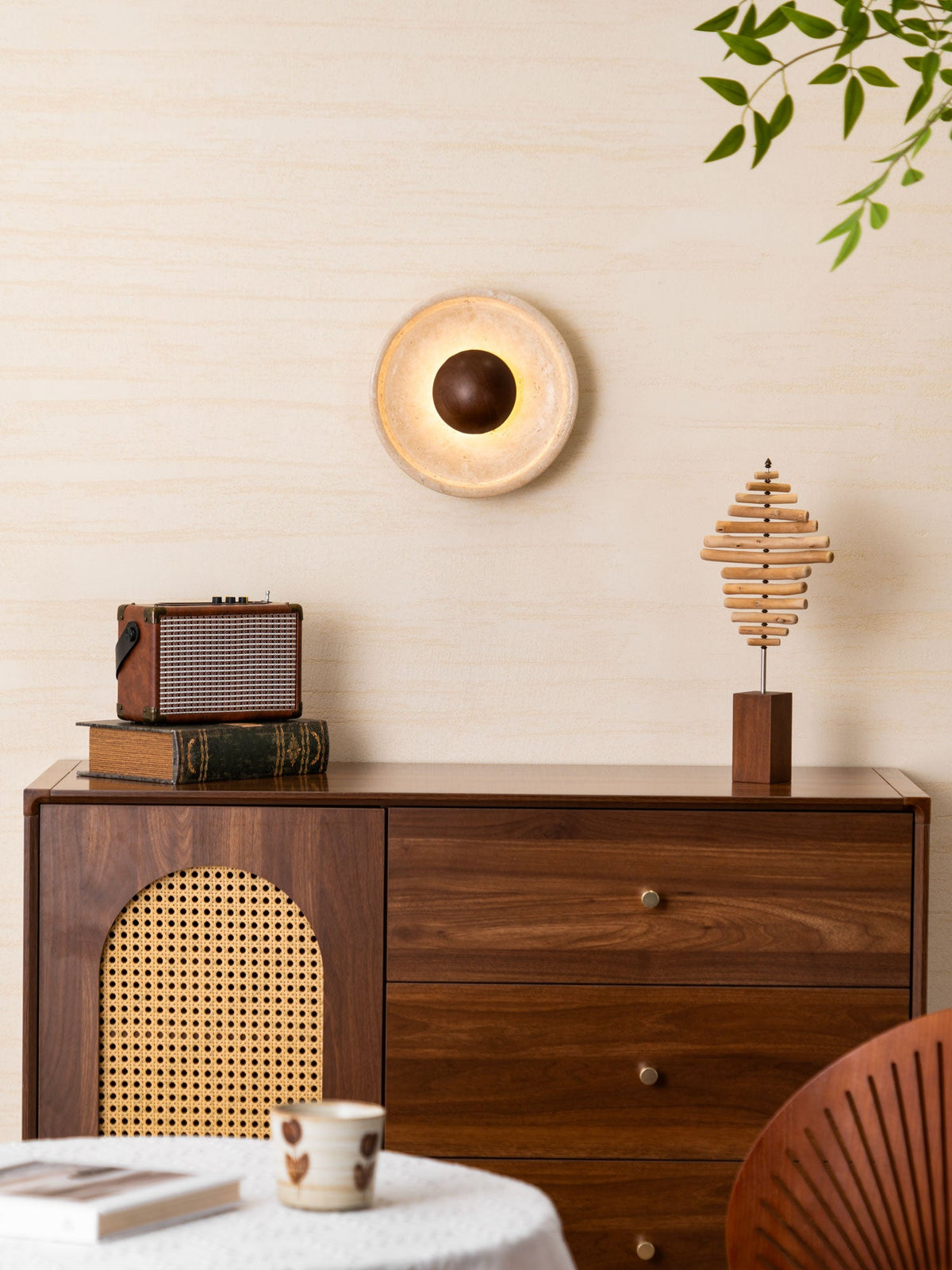This Clérachic wall light lit warm on a cream wall, above a walnut cabinet with a radio, a stepped wood sculpture and a cup.