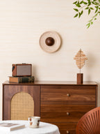 Unlit Clérachic travertine wall light with a walnut dome on a cream wall, above a walnut cabinet and green leaves at the top corner.