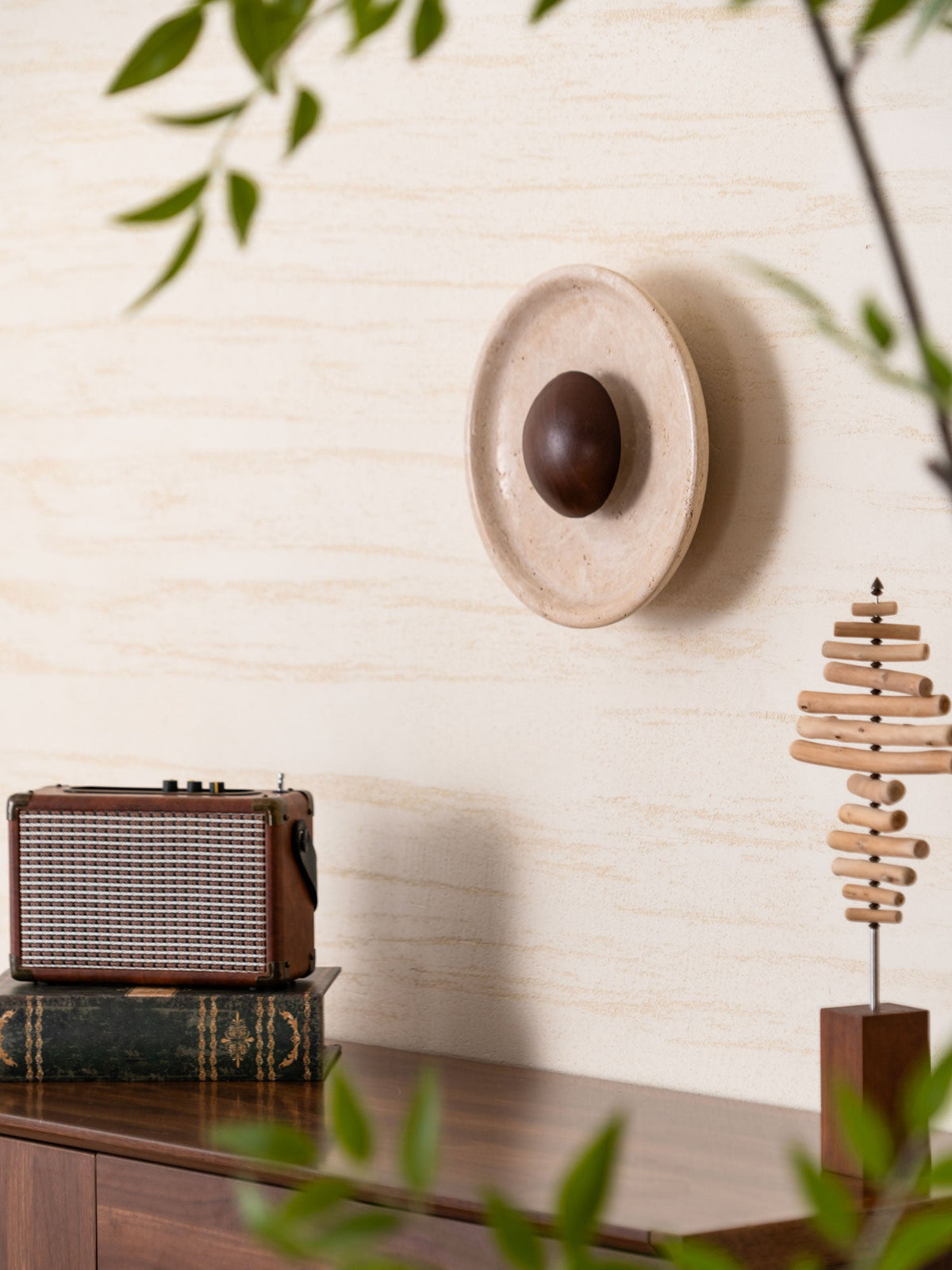 Angled view of the Clérachic travertine wall light with its walnut dome standing proud of the wall, with leaves in the foreground.