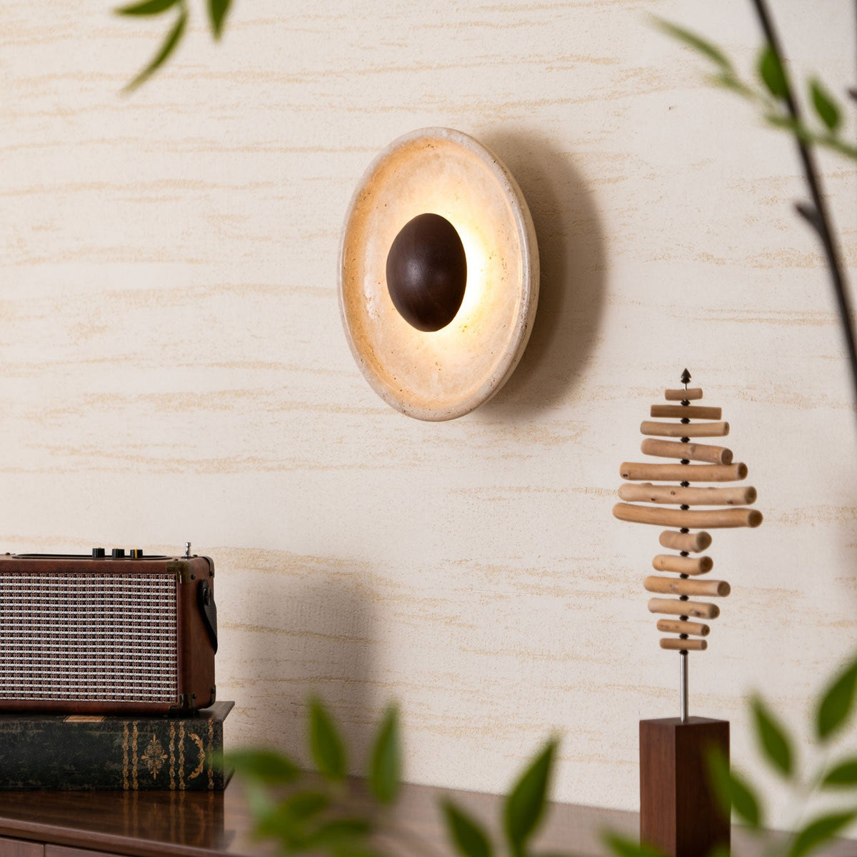 Side-on view of a lit Clérachic travertine wall light, with its walnut dome and a glow around the stone, near a radio and wood sculpture.