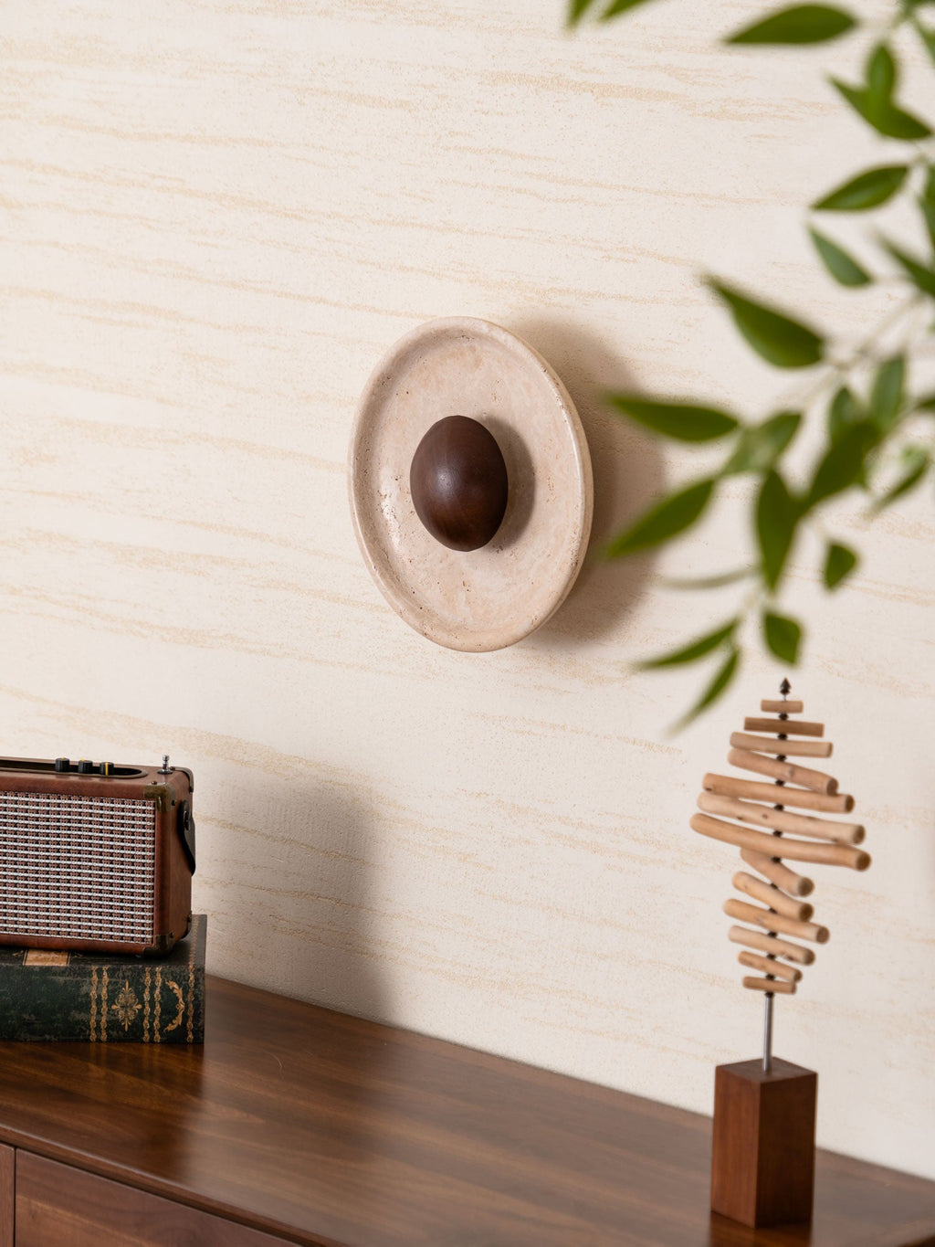 Seen from the side, the unlit Clérachic travertine wall light shows its raised stone rim and walnut dome, with green leaves in front.