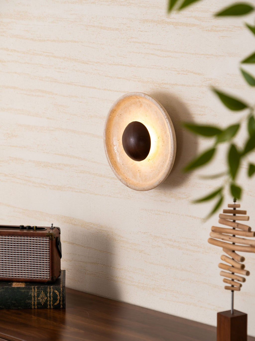 A lit Clérachic travertine wall light turned at an angle on a cream wall, the walnut dome ringed by warm light and framed by leaves.