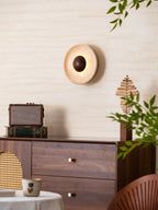 Clérachic wall light in travertine and walnut, lit, above a walnut cabinet with brass knobs, with foliage crossing the lower right corner.