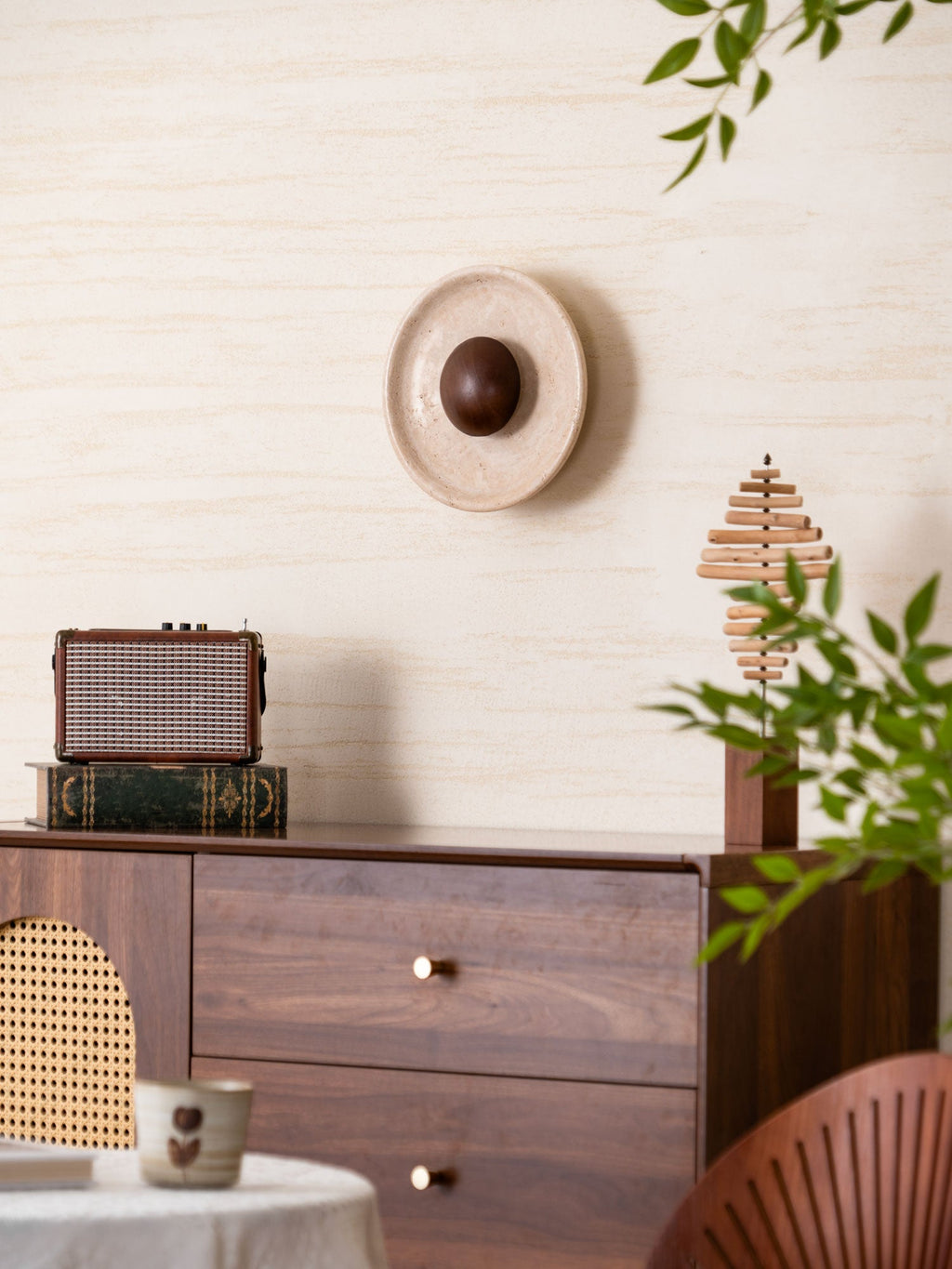 Unlit Clérachic travertine wall light with a walnut dome above a walnut sideboard with brass knobs, leaves in the lower right.