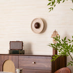 Clérachic travertine wall light with a walnut dome, off, on a cream wall above a radio and a wood sculpture, with leaves in front.