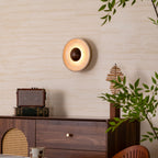 Lit Clérachic travertine wall light with a walnut dome, glowing softly above a walnut cabinet with a rattan door and leafy branches.